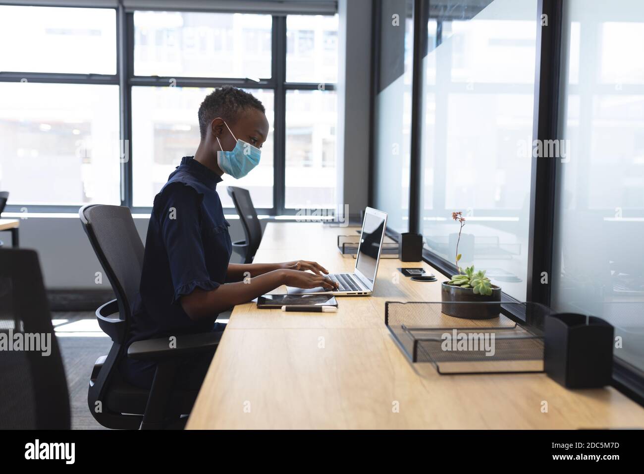 African american woman wearing face mask using laptop while sitting on her desk at modern office Stock Photo