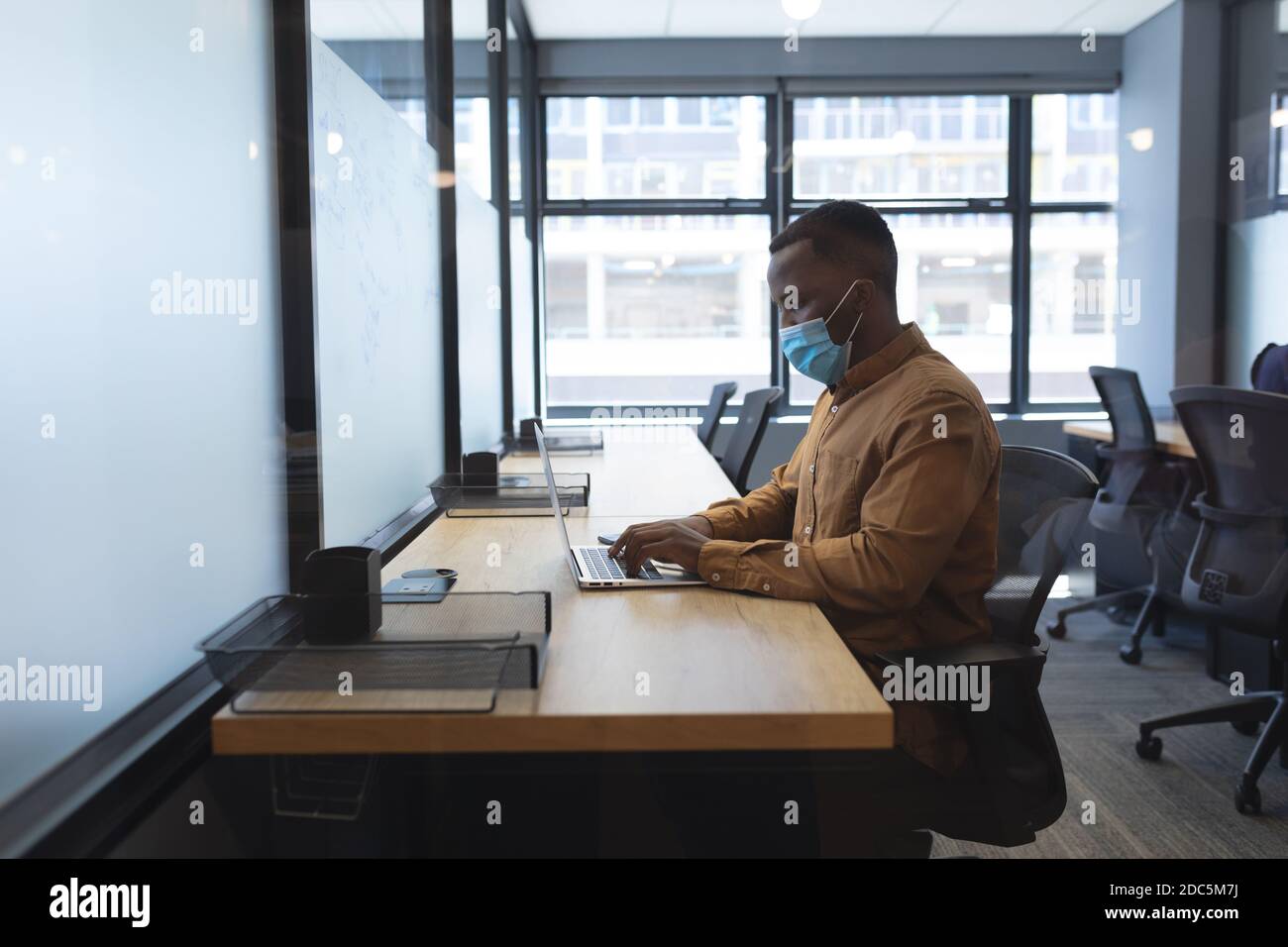 African american man wearing face mask using laptop while sitting on his desk at modern office Stock Photo