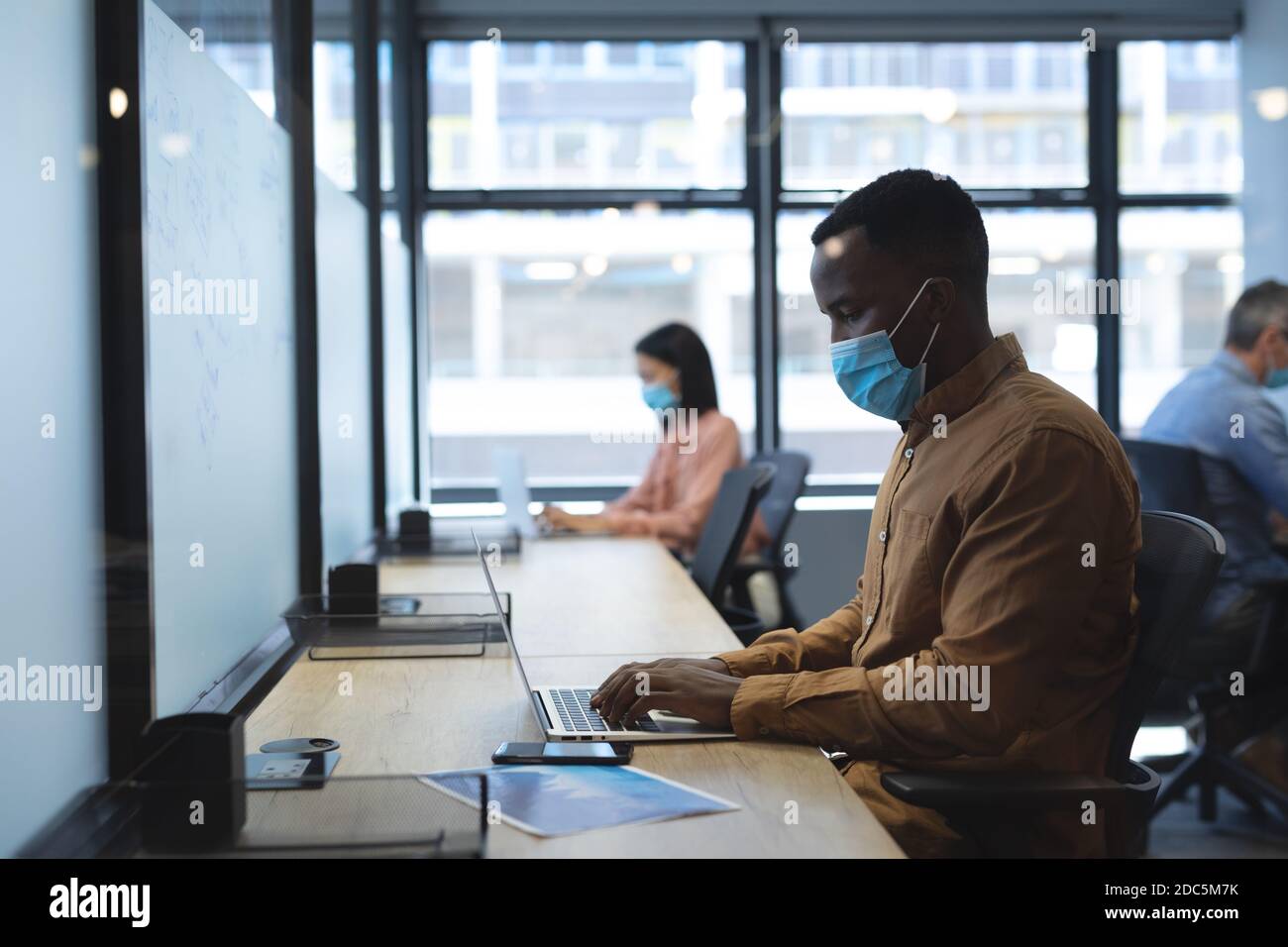African american man wearing face mask using laptop while sitting on his desk at modern office Stock Photo