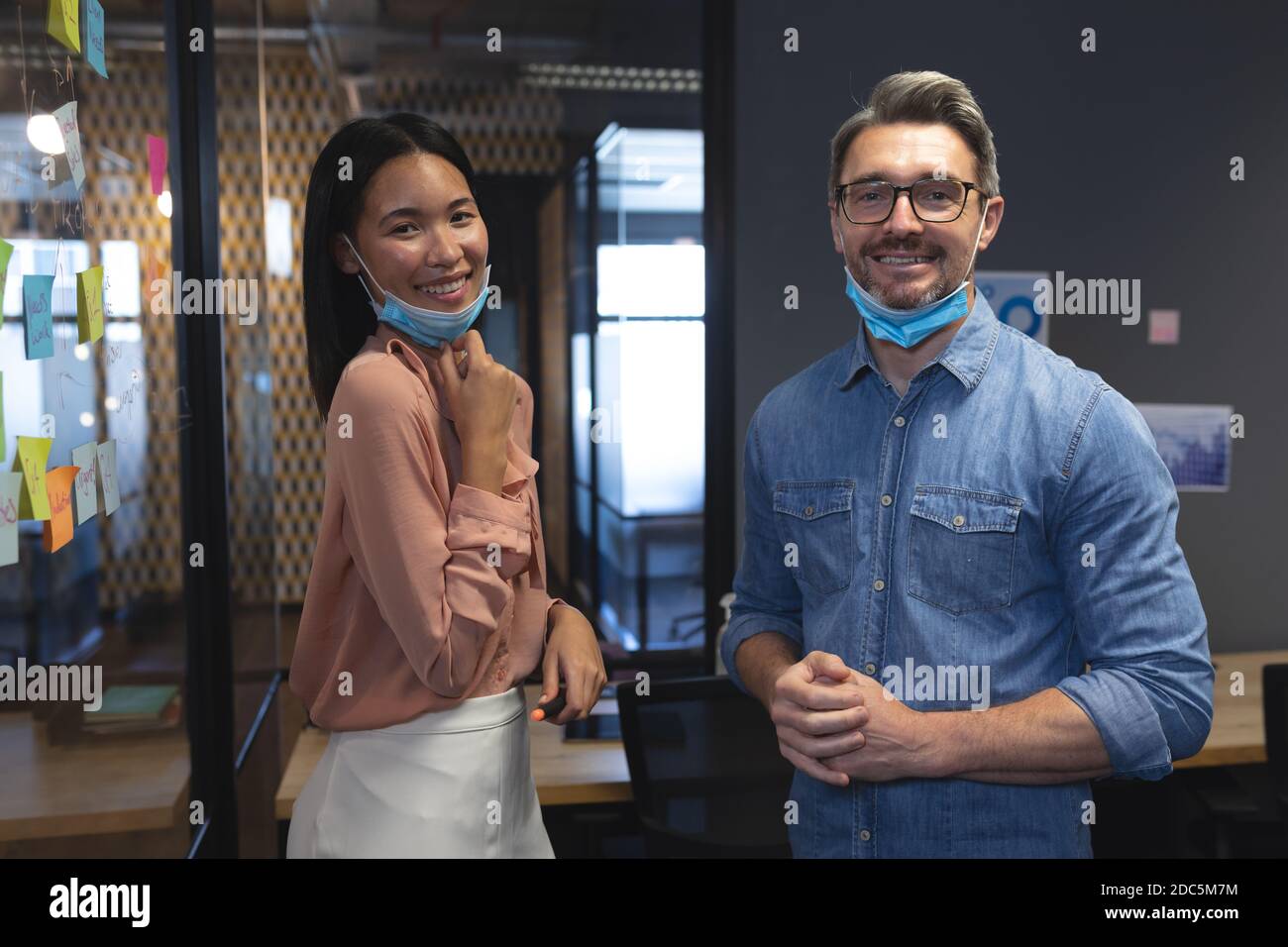 Portrait of caucasian man and asian woman with face masks around their neck smiling while standing i Stock Photo