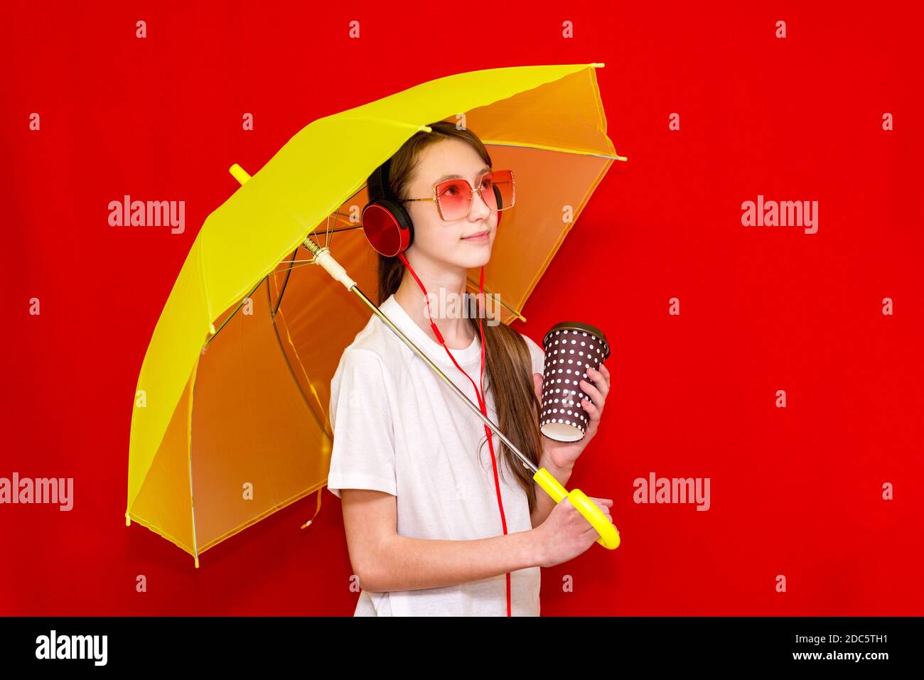 Teenage girl listens to music in headphones with umbrella. red studio background wearing white t shirt.studio shot. Stock Photo