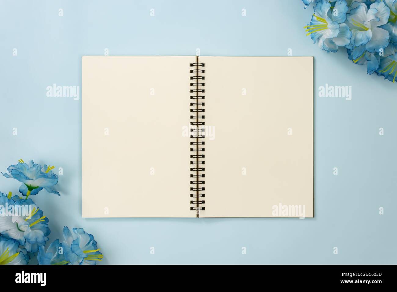 Top Table Spiral Notebook or Spring Notebook Mockup in Unlined Type on Center Frame and Blue Flowers at Bottom Left and Top Right Corner on Blue Paste Stock Photo