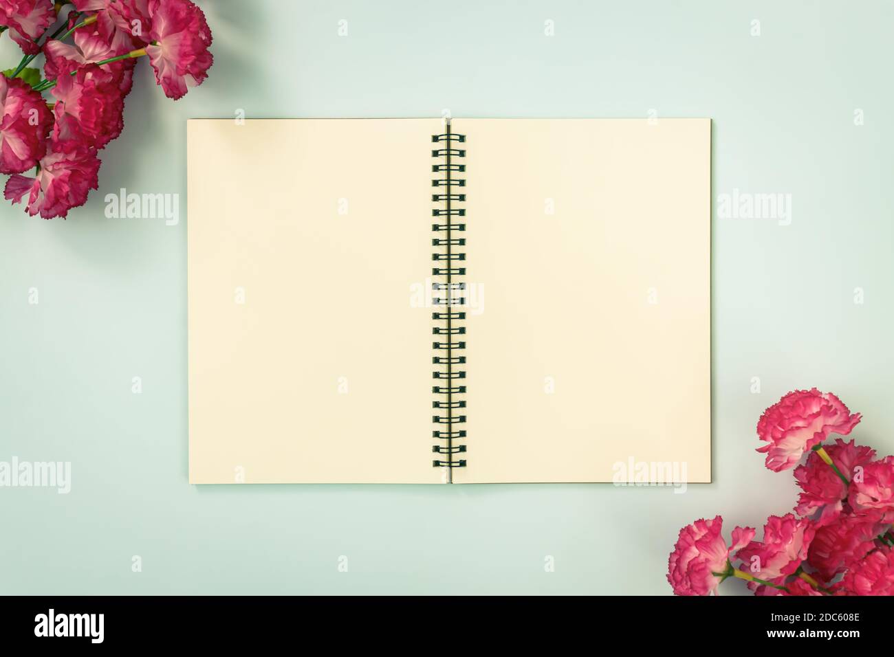 Top Table Spiral Notebook or Spring Notebook Mockup in Unlined Type on Center Frame and Red Flowers at Bottom Left and Top Right Corner on Blue Pastel Stock Photo