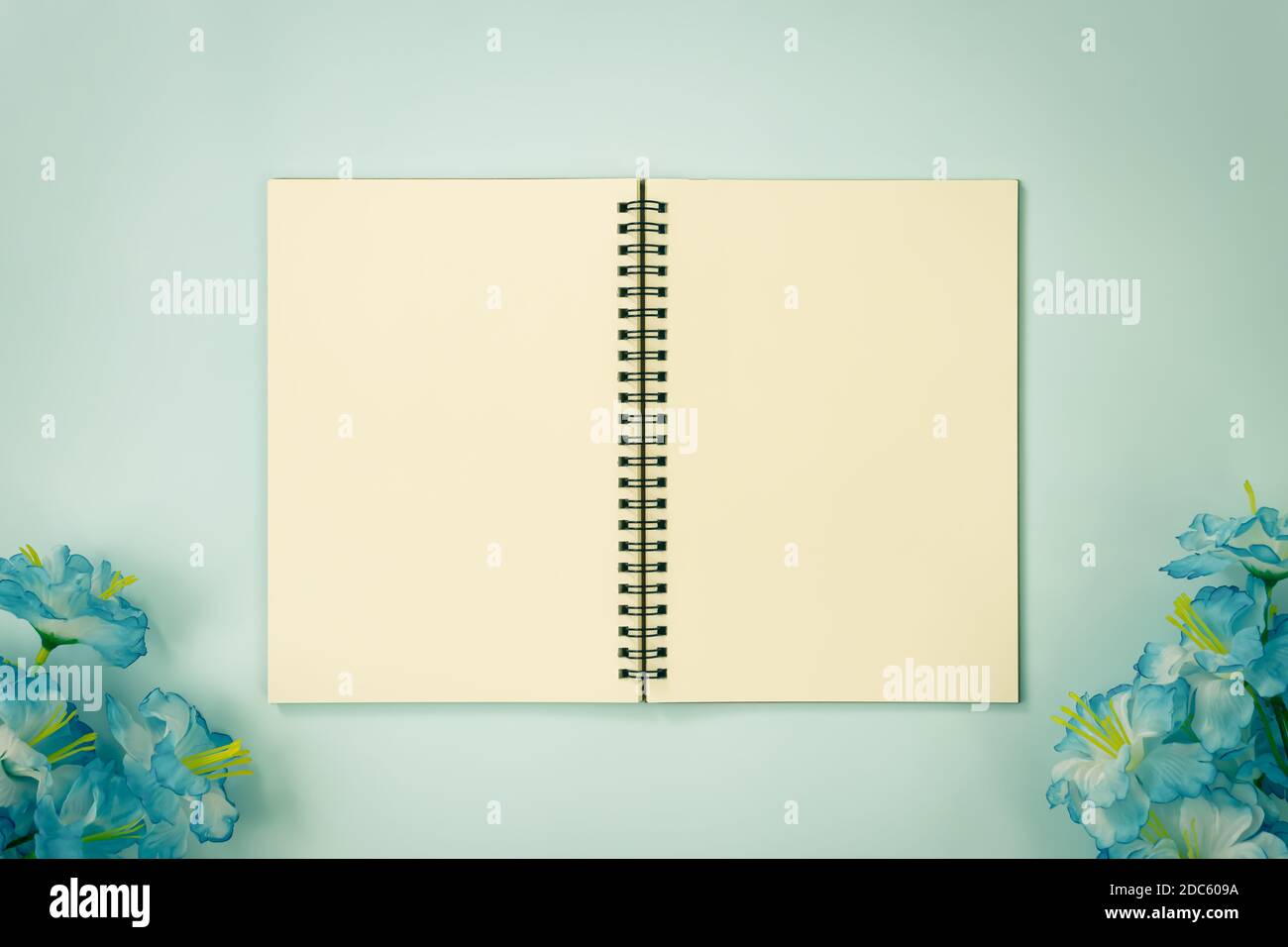 Top Table Spiral Notebook or Spring Notebook Mockup in Unlined Type on Center Frame and Blue Flowers at Bottom on Blue Pastel Minimalist Background in Stock Photo
