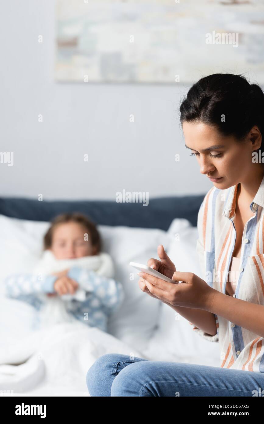 mother messaging on mobile phone near diseased daughter lying in bed on