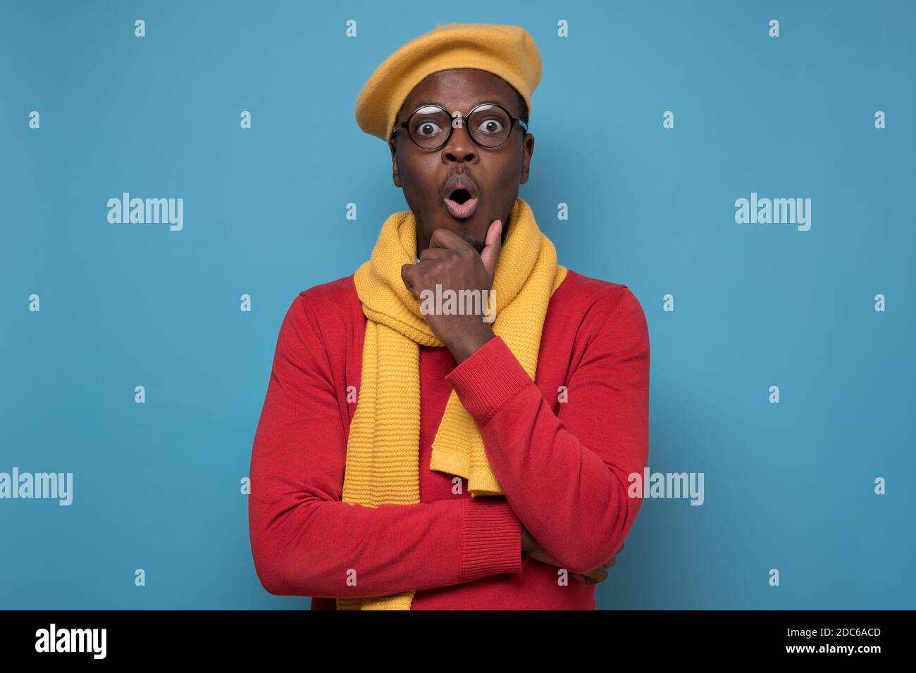 Young African American man wearing glasses and hat looking surprised. Studio shot on blue wall. Stock Photo