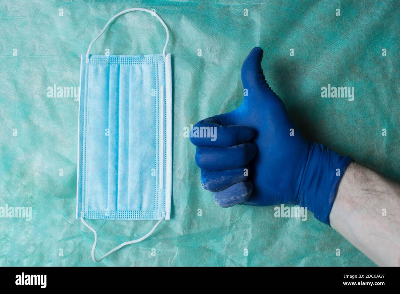A hand with thumb up as an approval gesture with a blue glove next to a surgical mask. Health care and surgical concept. Personal protective equipment. Stock Photo