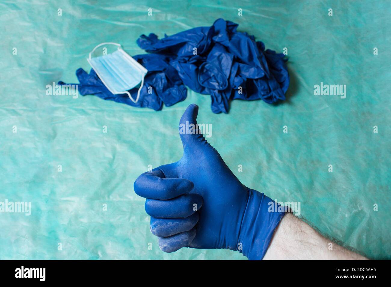 A hand with thumb up as an approval gesture with an unfocused pile of personal care equipment worn out in healthcare placed on green table. Stock Photo