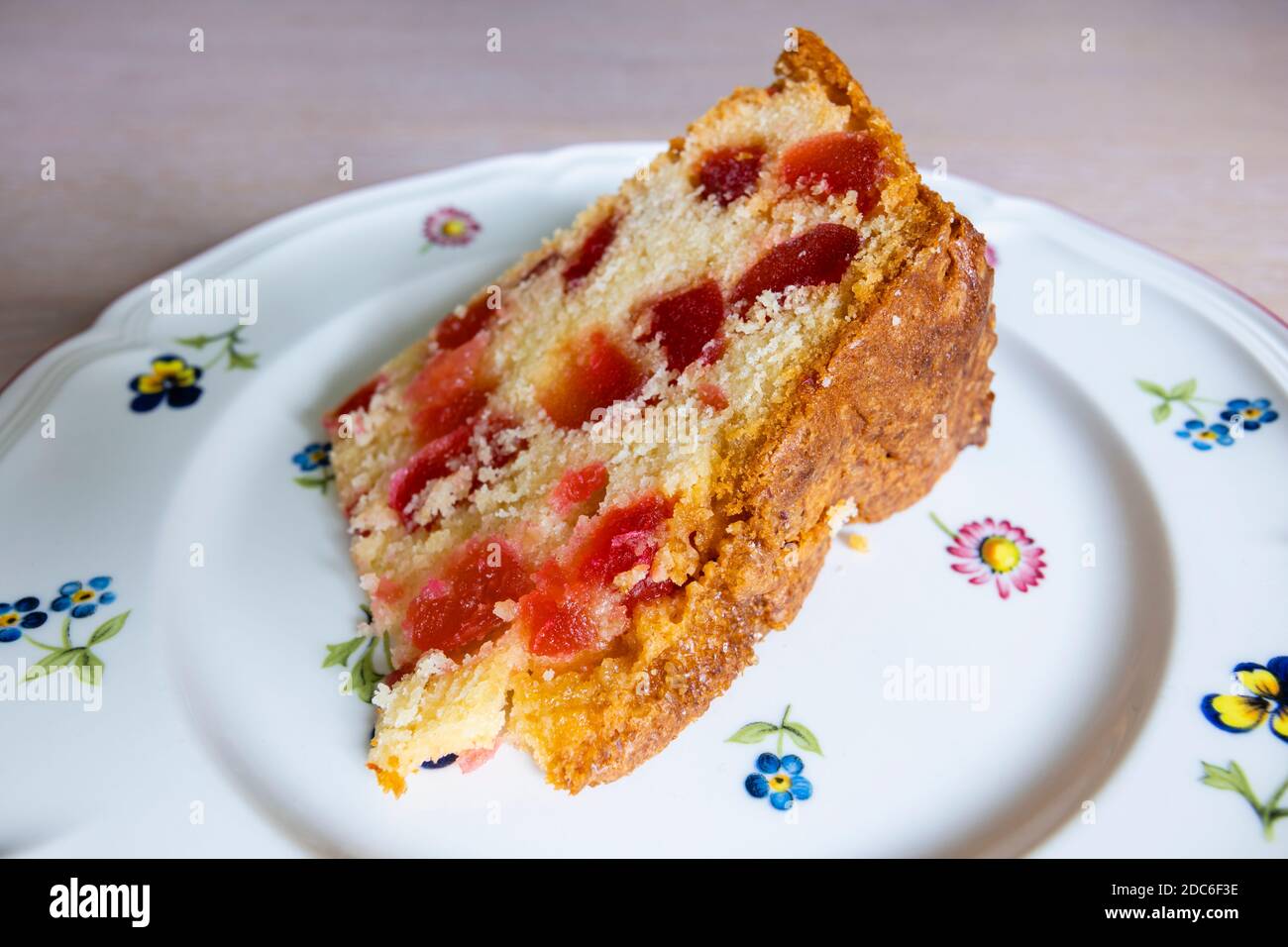 A cut slice of traditional round home-made cherry cake with red glace cherries served on a flowery white plate Stock Photo