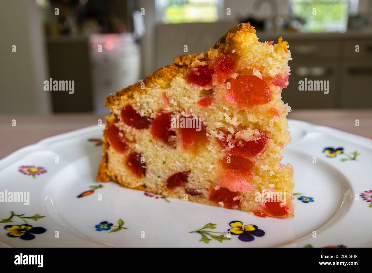 A cut slice of traditional round home-made cherry cake with red glace cherries served on a flowery white plate Stock Photo