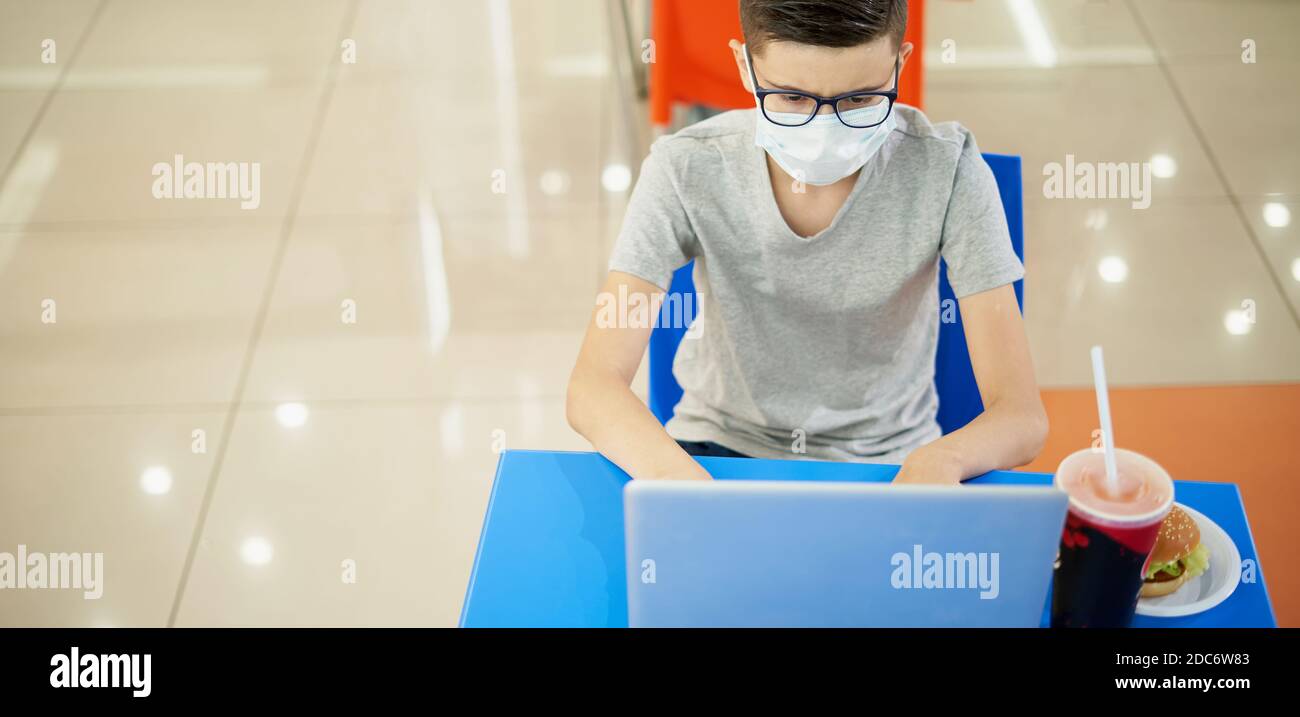 Boy teenager focused looking into laptop while sitting in food court of shopping mall. Boy in protective mask with laptop Stock Photo