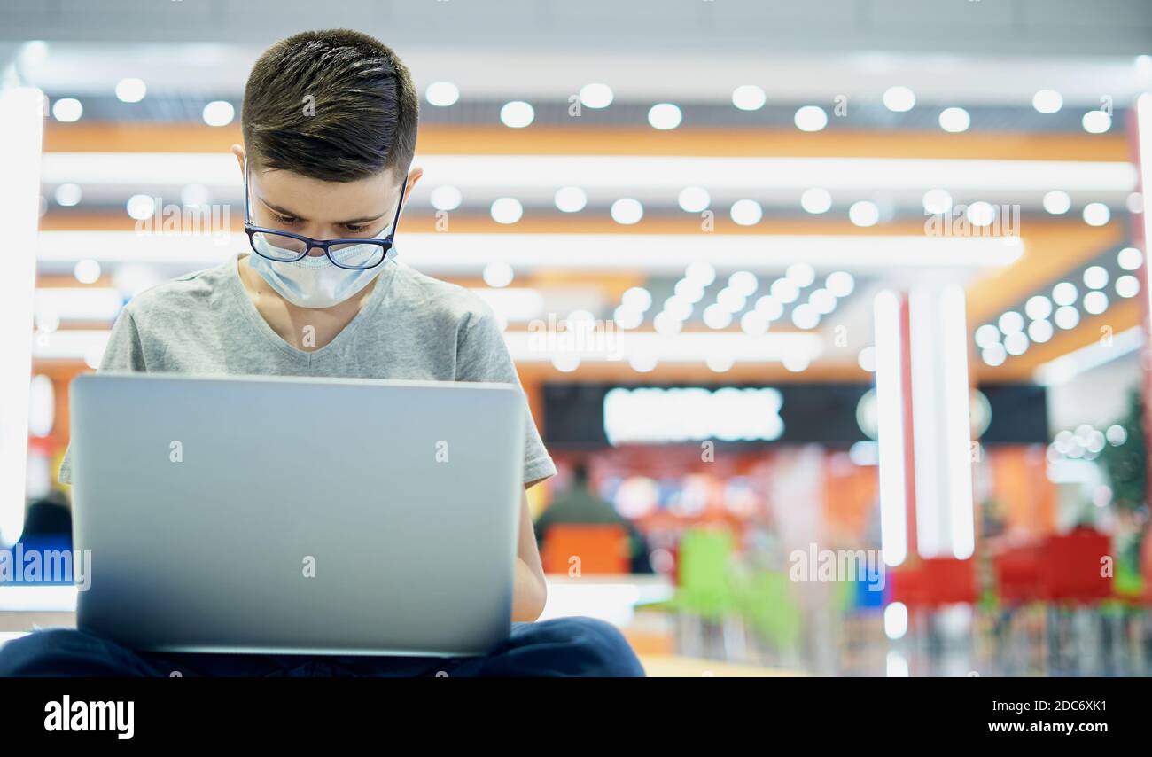 Teenager boy in protective mask uses laptop in shopping mall Stock Photo