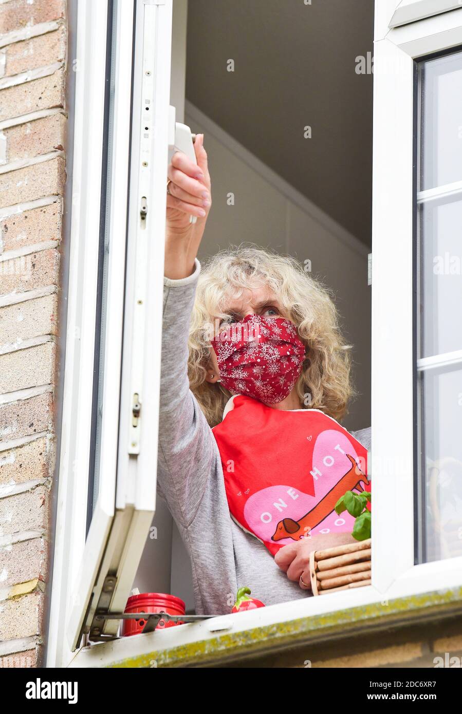 Woman holding a hot water bottle and wearing a face covering at home opening kitchen window to let in fresh air as advised to help combat coronavirus Stock Photo