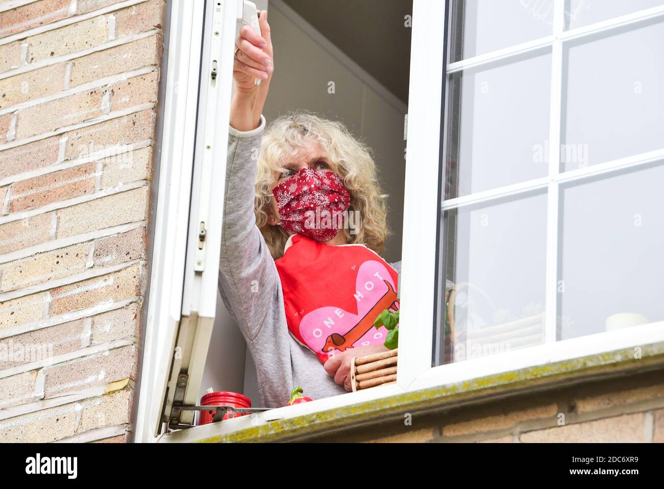 Woman holding a hot water bottle and wearing a face covering at home opening kitchen window to let in fresh air as advised to help combat coronavirus Stock Photo