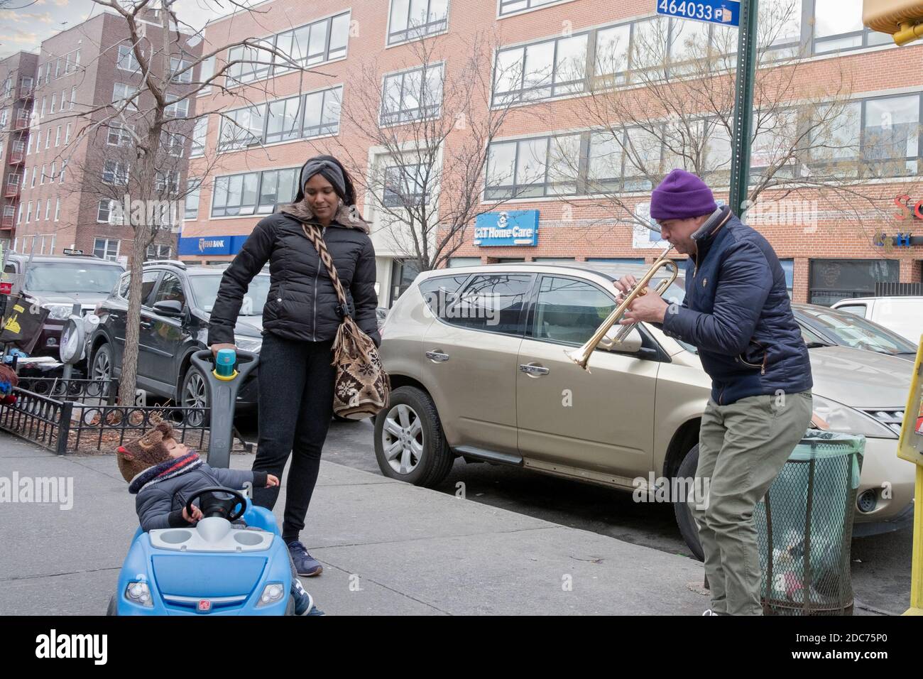 A trumpet playing street musician plays for a child and his mother. On 37th Avenue in Jackson Heights, Queens, New York City. Stock Photo