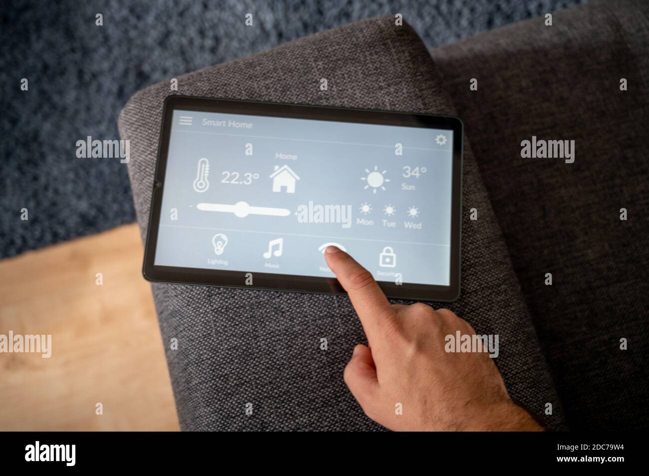 modern smart home control app with flat interface on black tablet display lies on the back of a sofa in front of a blue carpet a man controls the temp Stock Photo