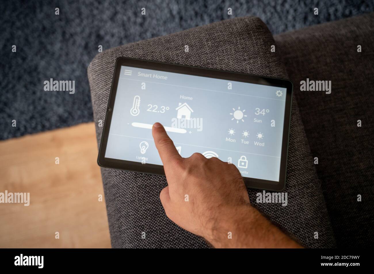 modern smart home control app with flat interface on black tablet display lies on the back of a sofa in front of a blue carpet a man controls the temp Stock Photo