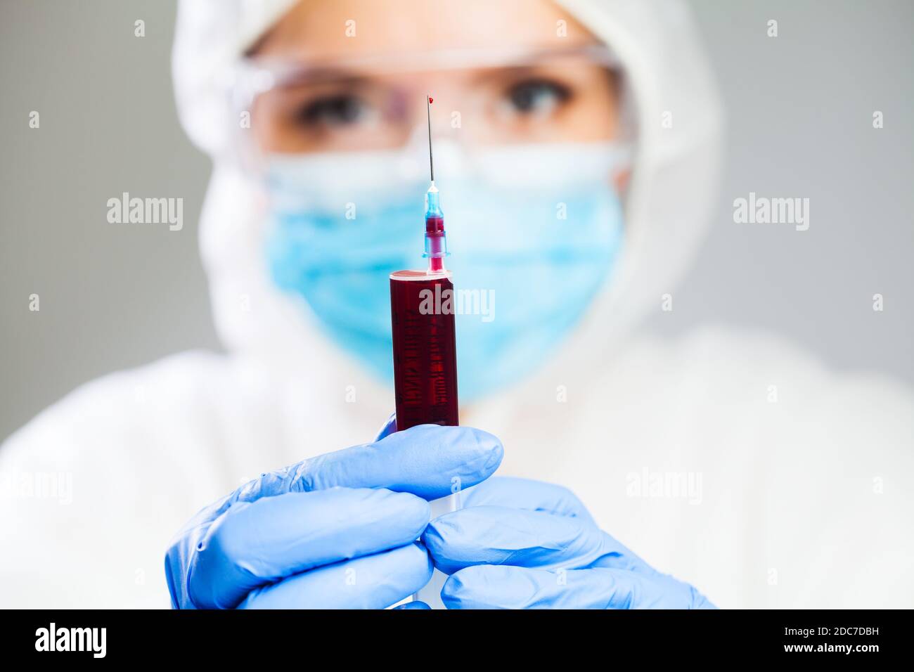Doctor or lab scientist in personal protective equipment holding syringe jab full of blood plasma,focus on single drop dripping out of the needle tip Stock Photo