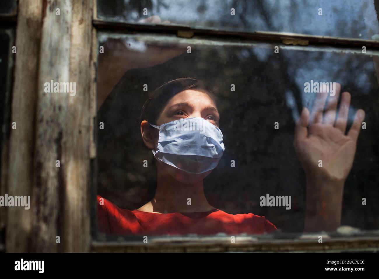 Sad depressed anxious young woman wearing protective medical mask looking outside the window, Coronavirus Covid-19 virus disease pandemic Stock Photo