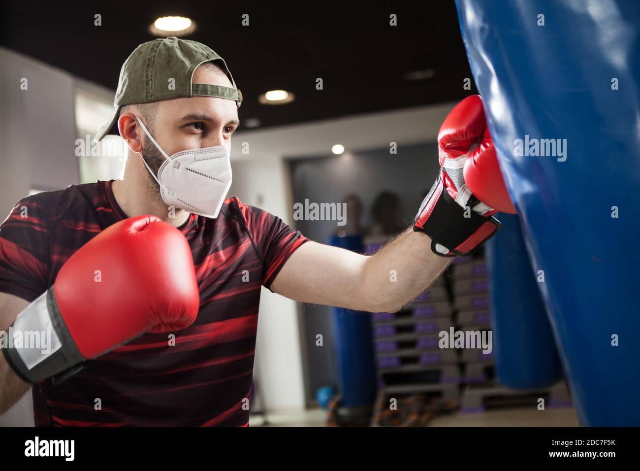 Young caucasian male wearing red boxing gloves & protective face mask,hitting blue sack in a reopen indoor gym,Coronavirus COVID-19 pandemic imposing Stock Photo