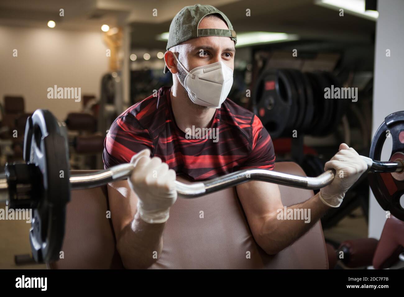 Young caucasian man working out in an indoor gym, barbell bicep curl exercise, wearing protective face mask, sport & fitness during COVID-19 pandemic, Stock Photo