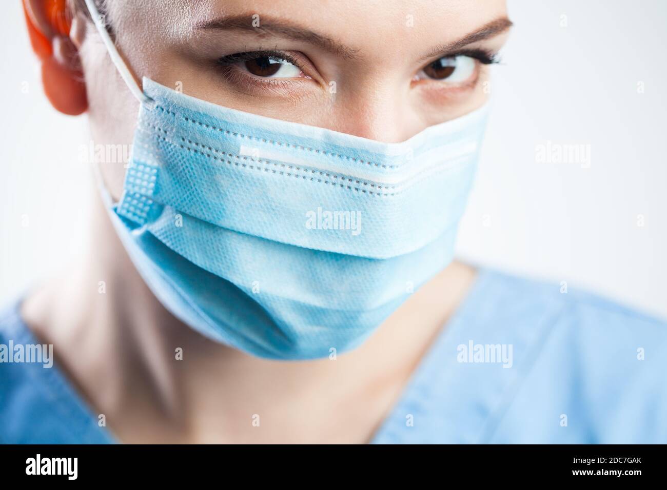 Young woman closeup detail headshot,wearing blue scrubs & protective face mask,attractive beautiful caucasian female doctor staring at camera Stock Photo