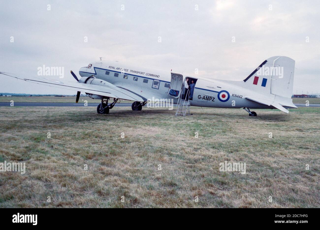 A 1999 photograph of a Douglas DC-3 Dakota, or C-47, aircraft, taken in England, registration G-AMPZ, painted in the colours of the British Royal Air Force Transport Command, with the military serial number KN442. The aircraft is parked on grass with two people visible in the doorway. Stock Photo