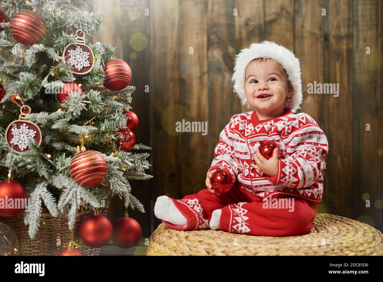 A happiness baby girl sits next to a Christmas tree and holds red Christmas balls Stock Photo