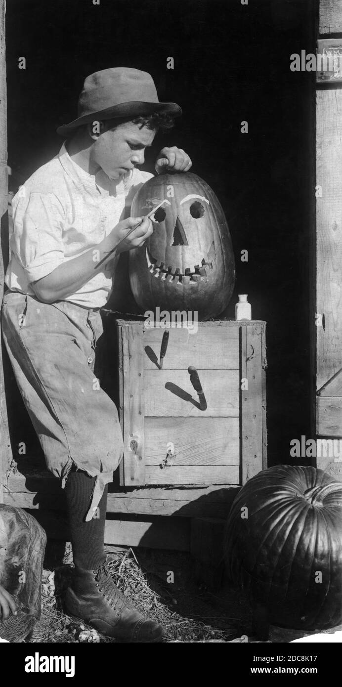 In the 1910s, a country boy has carved a large pumpkin into a Jack-o-Lantern with a big, toothy smile. He is now concentrating on painting white eyebrows onto the pumpkin's face. The pumpkin's box has two opened jackknives sticking into it, and along with the boy, it is all framed in a shed's doorway. At below right, a second pumpkin awaits its turn under the knife.   To see my other holiday images -- Search:  Prestor  vintage  holiday Stock Photo