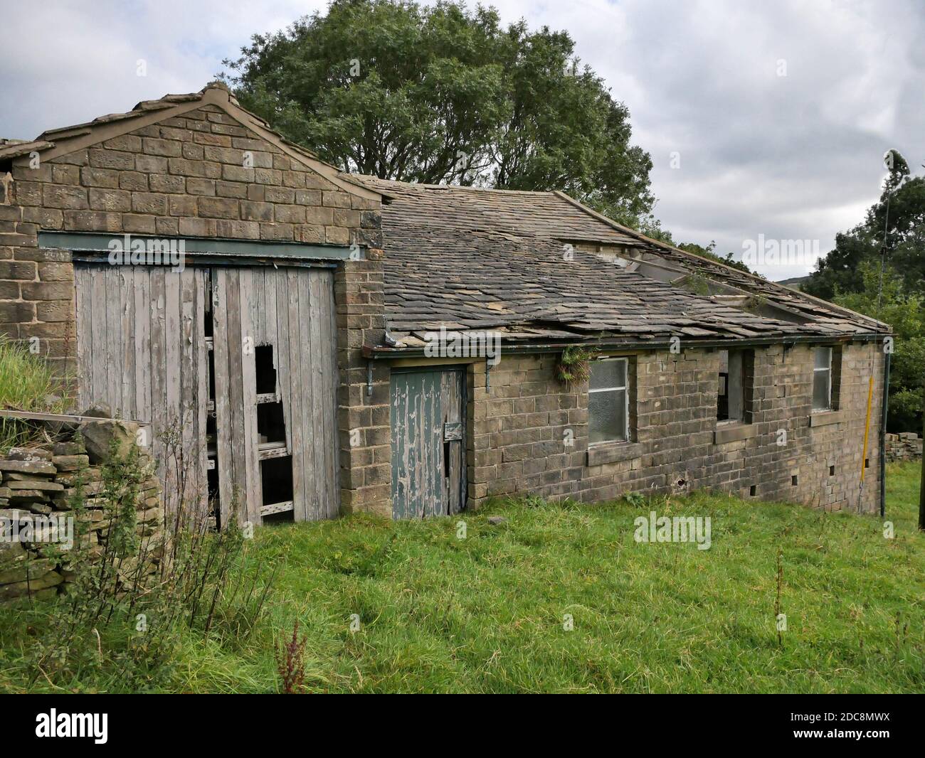 Abandoned old stone built single story barn with roof collapsed old wood doors falling apart field and trees in background Stock Photo