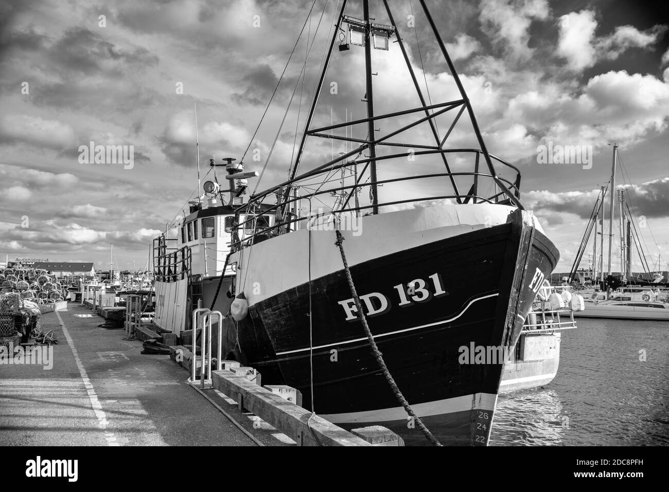 A trawler is moored alongside a wharf and the yachts of a marina are beyond.  A blue sky with clouds is above. Stock Photo