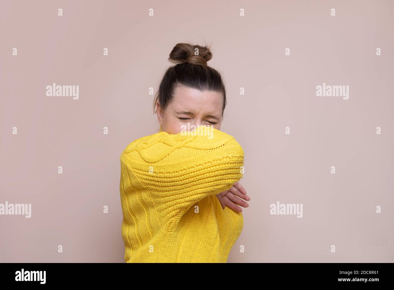 Photo of woman in a yellow sweater with a bun on her head on a pink background. Sneezes or coughs into the elbow. Safety concept, culture of behavior Stock Photo