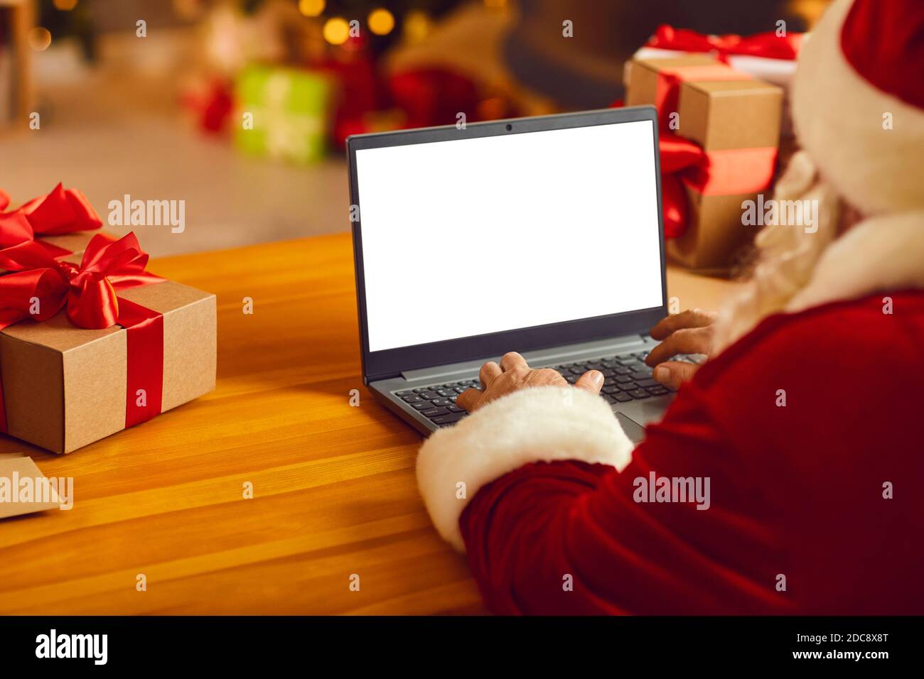 Senior Santa in festive traditional costume and hat sitting typing Christmas congratulation online on laptop Stock Photo