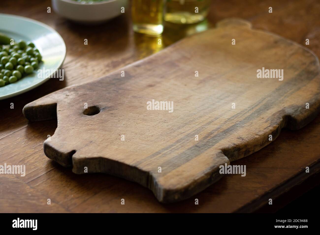 wooden cutting board shaped like a piggy, a plate with green peas and a bottle of oil on a wooden table Stock Photo