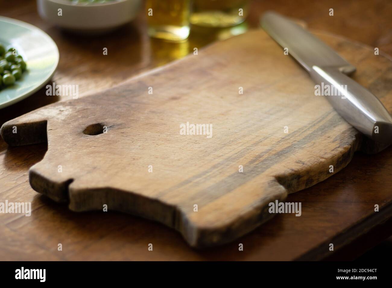 wooden cutting board shaped like a piggy with a kitchen knife, a plate with green peas and a bottle of oil on a wooden table Stock Photo