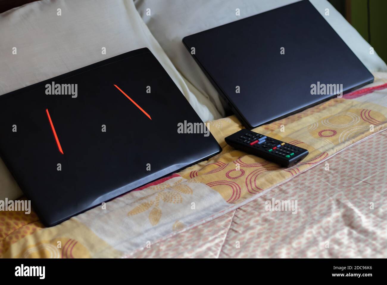 Two black laptops and a tv remote control on bed with pillows