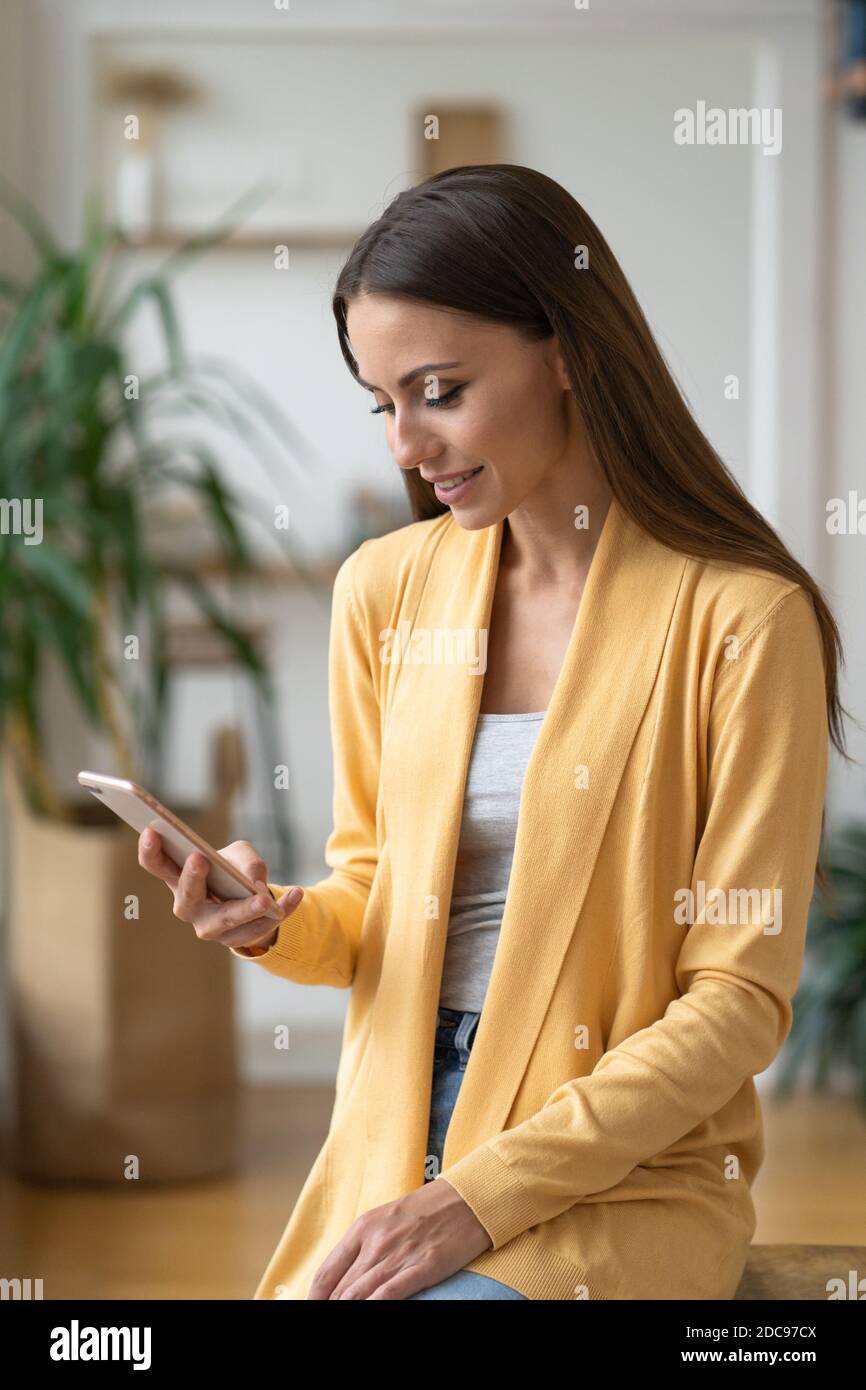 Satisfied cheerful woman wear yellow cardigan holding mobile phone, makes shopping online, surfing internet received message from friend chatting abou Stock Photo