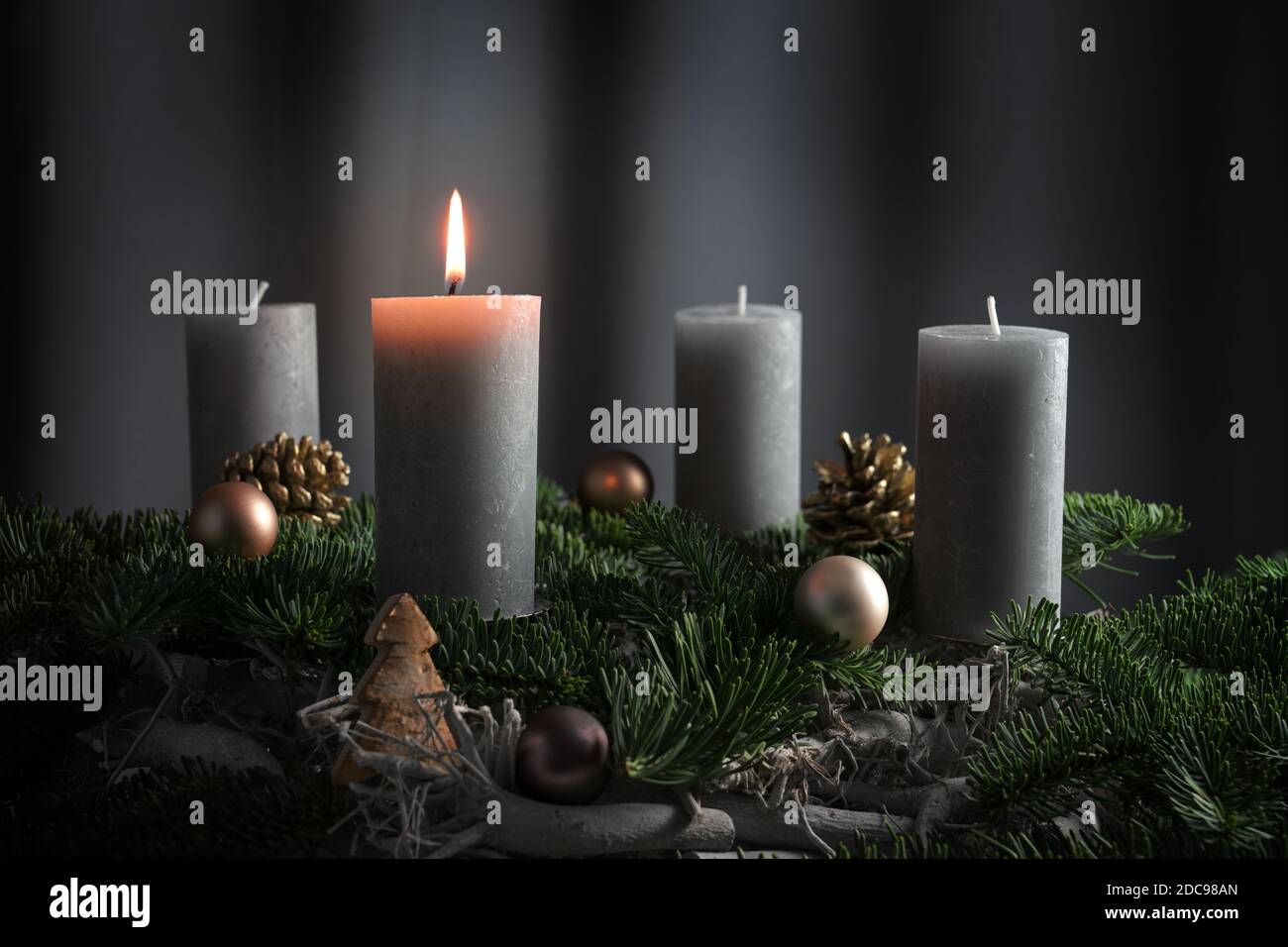 Four candles, one of them lit on an advent arrangement from fir branches and Christmas ornaments, holiday decoration against a dark gray background wi Stock Photo