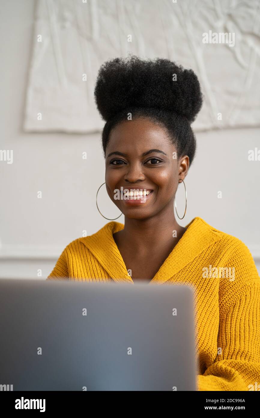 Cheerful African American biracial millennial woman with afro hairstyle in yellow cardigan remote working on laptop, looking at camera, has good mood. Stock Photo