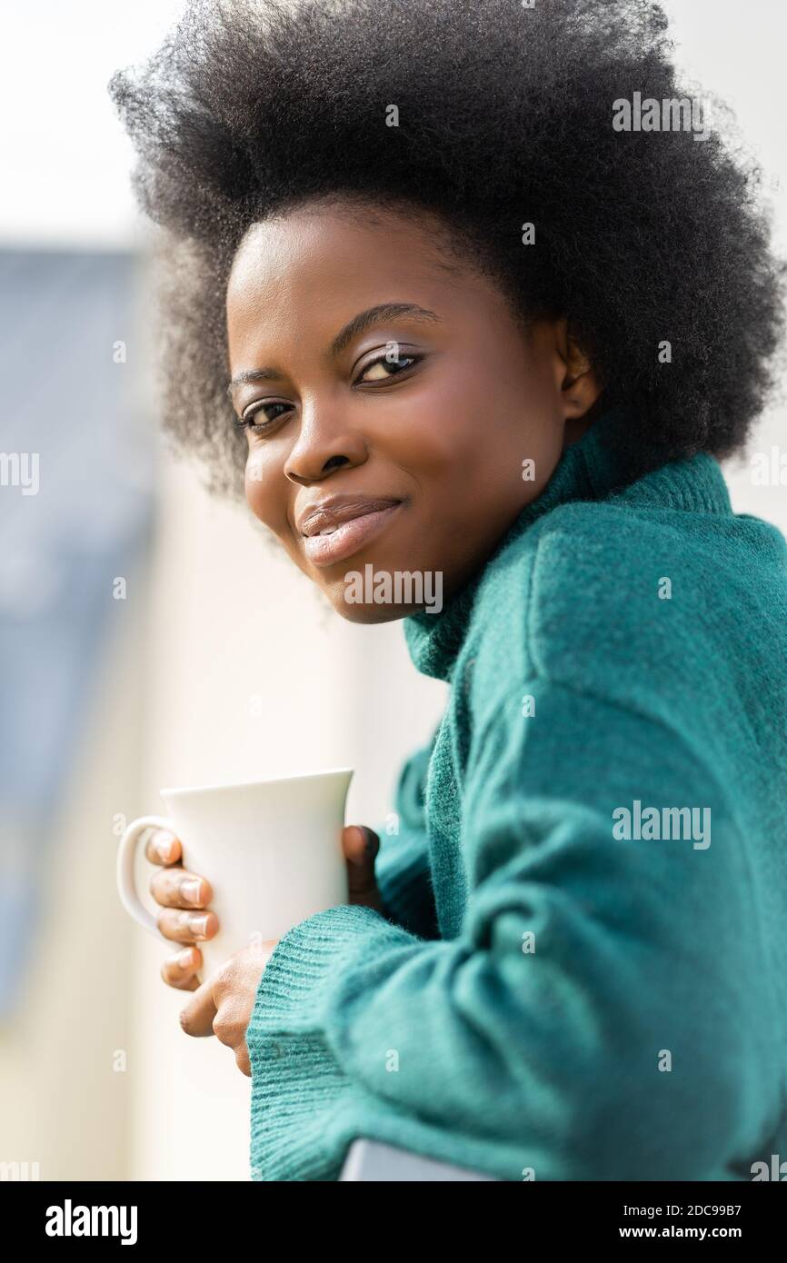Cute young African American biracial woman enjoying a cup of tea or coffee, wear oversize green knitted sweater, looking at camera, standing on the ba Stock Photo