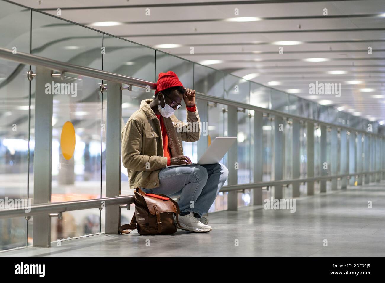 Stylish Black traveler millennial man wear face protective mask, sitting in empty airport terminal, works remotely on laptop while waiting for a fligh Stock Photo