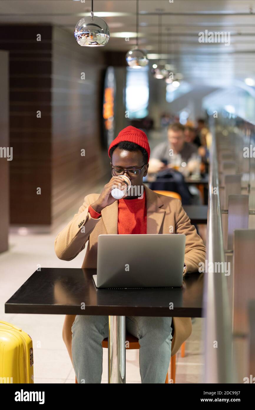 Afro-American traveler millennial man in beige coat, red hat sitting at cafe table in airport terminal, drinking coffee or tea, works remotely on lapt Stock Photo
