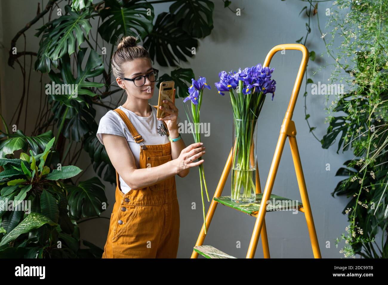 Smiling woman florist in orange overalls takes a photo of bouquet of purple irises in glass vase on smartphone, standing on stepladder. Love for flowe Stock Photo
