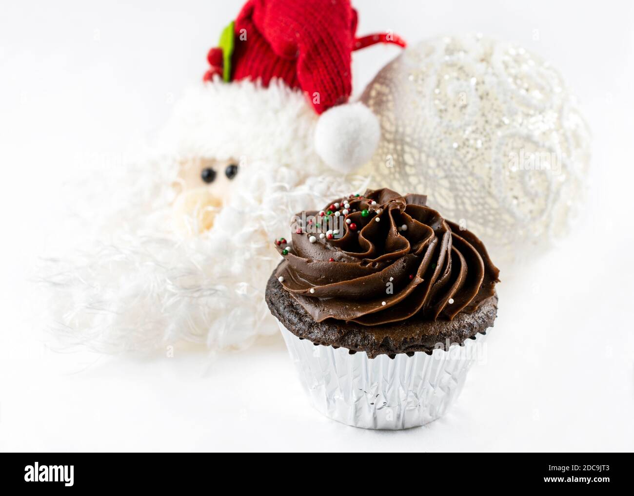 A close up shot of a Christmas chocolate cupcake with small candy sprinkles of red, green and white.  Santa and ball ornaments blurred in background. Stock Photo