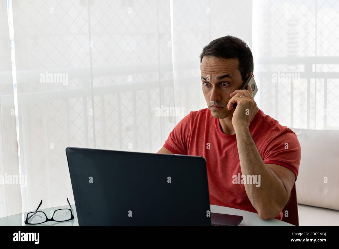Mature man with cell phone in hand, looking at laptop with wide eyes. Stock Photo