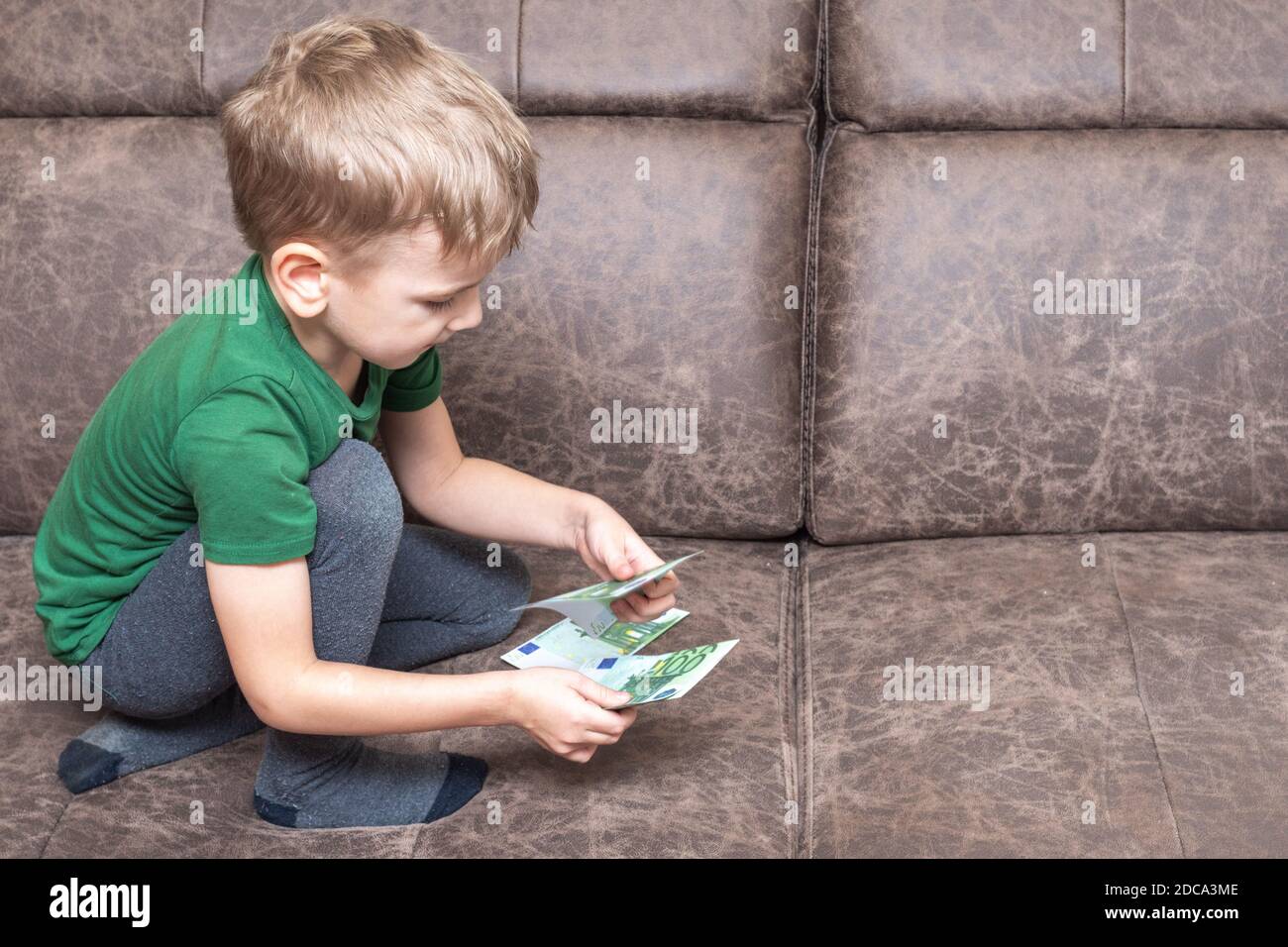 Concentrated caucasian child in casual clothes holds one hundred euros in his hands and counts money on the sofa in the room, side view, copy space. C Stock Photo