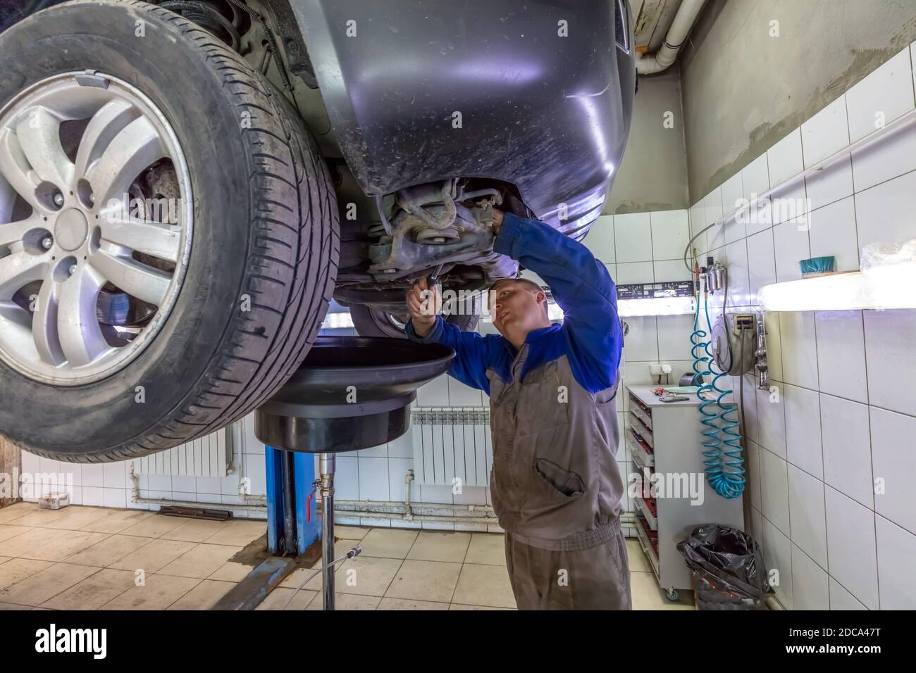 Car mechanic changes oil in a workshop. Mechanic standing under the car and draining oil into a special cannister via funnel. Stock Photo