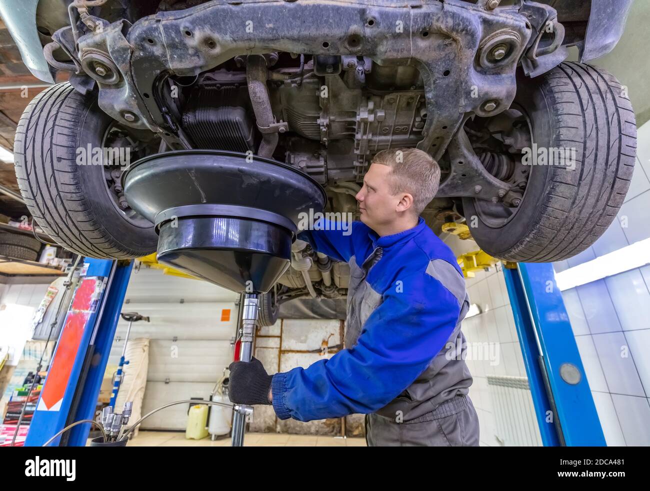 Happy car mechanic changes oil in a workshop. Mechanic standing under the car and draining oil into a special cannister via funnel. Stock Photo