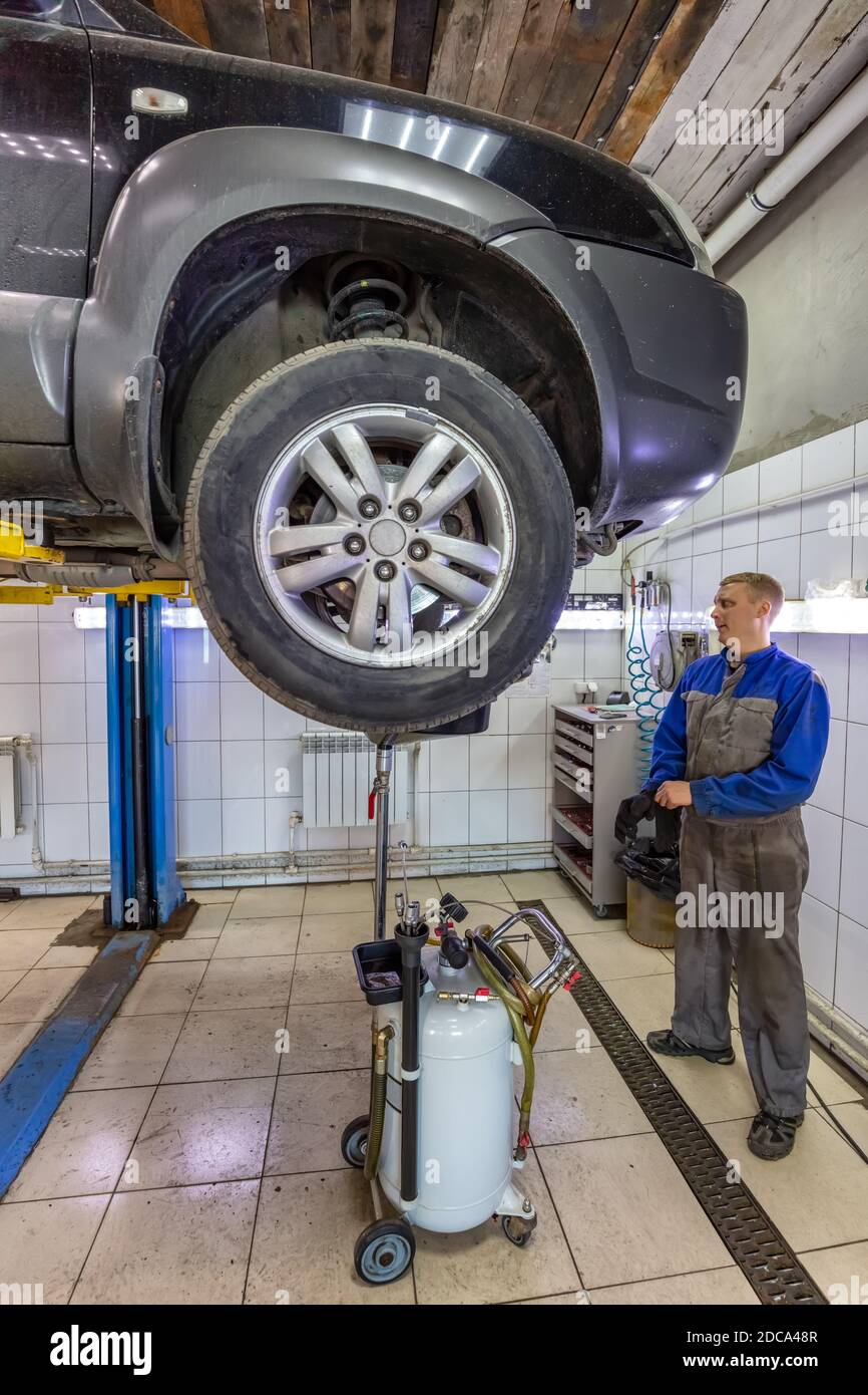 Car mechanic changes oil in a workshop. Mechanic standing next to the car and draining oil into a special cannister. Car is on the hydraulic lift. Stock Photo
