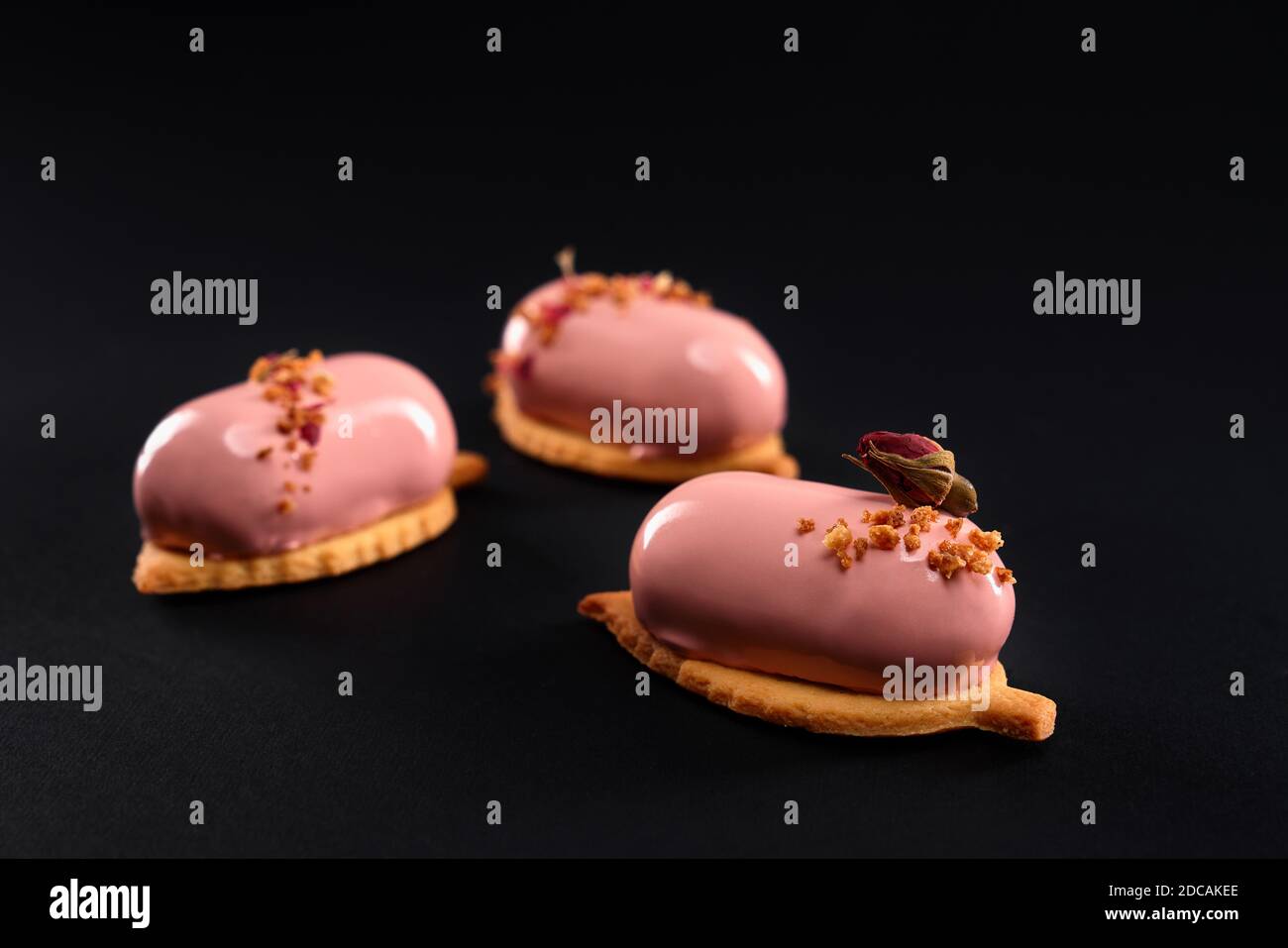 Pink cakes decorated with dry rose flower and nuts. Dessert with smooth surface, mousse and mirror glaze on cookie isolated on black studio background. Tasty sweet dish in cafeteria. Stock Photo